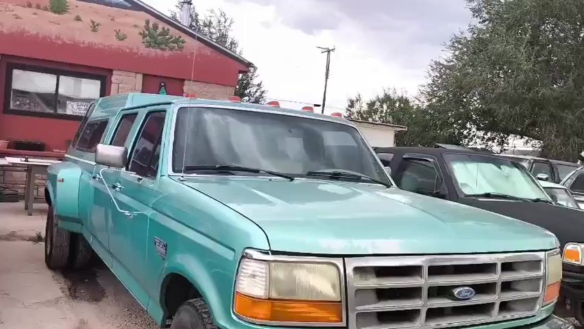 The video pans across a car repair shop and parking lot. The building has a large mural on its side featuring characters from the movie Cars and a Route 66 sign. There are several vintage cars in various states of repair parked in front of the building, including a black Mustang with its hood open, a black and white two-tone car on jack stands, and a teal pickup truck. To the left, there's a building with a large American flag mural and several white vehicles, including a van and an SUV. The sky is overcast.
