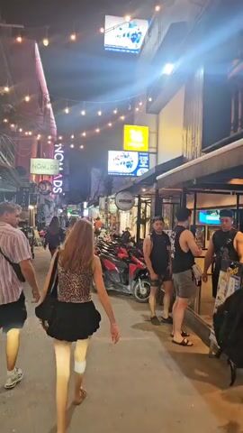 The video depicts a bustling street scene at night in Ko Tao, Thailand. String lights crisscross overhead, illuminating the narrow thoroughfare lined with various shops and bars. Numerous people are seen walking along the street in both directions, enjoying the evening atmosphere.

In the foreground, a woman wearing a leopard-print top and a black skirt walks away from the camera, accompanied by a man in a striped shirt. Further down the street, other individuals and small groups stroll past storefronts. A man in a black tank top with "KOH TAO" printed on it and shorts is visible near the entrance of a bar, where glowing green screens can be seen inside. Another man, wearing a light-colored shirt and sunglasses, is seated on a black scooter with the number "15" on its front, briefly looking towards the camera before turning his head. Several other scooters are parked along the side of the road.

Illuminated signs from businesses are visible, including a prominent yellow sign displaying the "SINGHA" lion logo and another blue sign above it. To the left, a sign reads "NOMADS." The scene suggests a lively commercial or entertainment district, with people casually walking and socializing.