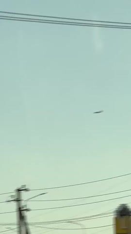 A low angle shot shows a pale blue sky with a faint contrail. In the distance, a small airplane is visible. In the foreground, a concrete pillar stands on the left, and power lines and a utility pole are visible across the bottom of the frame.