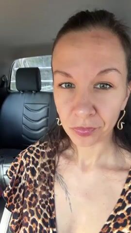The video shows two women in a car and a man outside the car. The woman in the foreground is looking at the camera. She is wearing a leopard print top. The woman in the background is looking out the window. She is wearing a red and white floral top and blue jeans. A bottle of water and a purse are on the center console of the car. A man is standing outside the car window, holding a green basket filled with various items. He appears to be interacting with the people inside the car. The car is parked on the side of a road, with a rural landscape visible outside the windows.