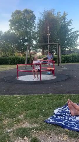 Two children are on a playground. One child is sitting on a merry-go-round and the other child is standing and appears to be pushing the merry-go-round. The children are wearing casual clothing. There is a swing set in the background, along with trees and bushes. The ground is covered in a dark surface with some grass in the foreground. A person's feet are visible in the bottom right corner of the frame, lying on a blanket. The lighting suggests it is daytime, possibly late afternoon.