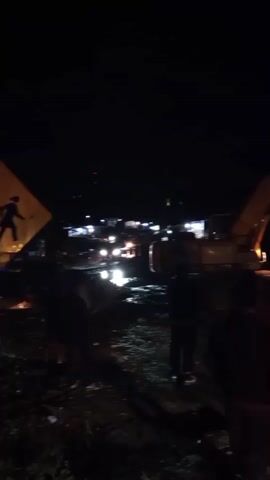 The video is dark and grainy, suggesting it was recorded at night with poor lighting. It appears to capture an outdoor scene, possibly a construction site or an area with ongoing work. Several people are visible in the foreground, mostly silhouetted against the dim light. In the background, there is a large yellow excavator with its arm raised, illuminated by its own lights. Other lights, possibly from vehicles or buildings, are also visible in the distance. The ground appears to be uneven, with debris or rough terrain. There is a sense of activity, with people gathered and heavy machinery present. The overall atmosphere is dim and somewhat obscured due to the low light conditions.