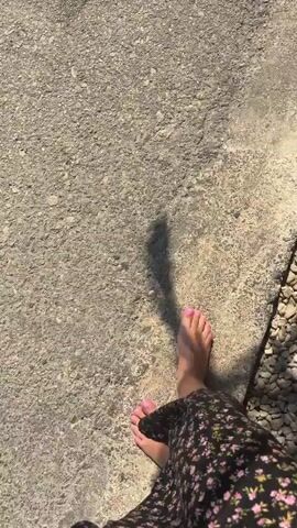 A person is walking barefoot on a rough, stone path toward the ocean. They are wearing a dark floral print dress. As they approach the water, the waves lap at their feet. The scene is shot from a first-person perspective, looking down at the person's feet and legs.