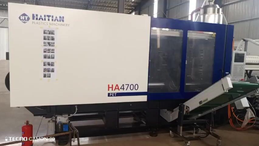 The video shows a large, industrial Haitian Plastics Machinery HA4700 PET injection molding machine. To the right of the machine, there is a conveyor belt leading away from it. Behind the machine are other pieces of industrial equipment, including a ladder propped against the wall in the background. To the right of the equipment are stacks of large, white bags tied with green string, some labeled "Abril Agro" and "MAMCO". The scene is inside a large, corrugated metal industrial building.