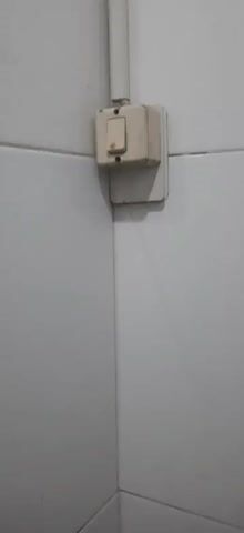 The video shows a close-up view of a corner where two white tiled walls meet. A white electrical conduit runs vertically down the corner, attached to the wall. Near the bottom of the conduit, a beige electrical switchbox is mounted. The switchbox has a single toggle switch that appears to be in the "off" position. The conduit continues upwards, disappearing from view. Further up the corner, a brown trim piece runs horizontally along the top of the tiled wall. Above this trim piece, a square white exhaust fan is visible, recessed into the ceiling. The fan has a circular grille with concentric rings. The overall environment appears to be an interior space, possibly a bathroom or kitchen, given the tiled walls and exhaust fan. The lighting is somewhat dim, casting faint shadows. There are no people or animals visible in the video.