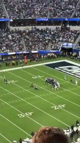 The video shows a wide, high-angle view of an American football game in a large stadium packed with spectators. The teams, one in white and the other in black uniforms, are positioned on the field near the end zone with the word “CHARGERS” displayed in large letters. A player in white kicks the football, and the ball goes through the goalposts. The crowd reacts with cheers and applause. The field is green with yard line markings. The environment suggests a well-attended professional sports event.