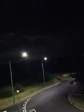 The video shows a dark, unlit rural road at night. There are two streetlights visible, one brighter than the other. The road curves to the right. The sky is dark with some clouds. There is grass and vegetation on the sides of the road. No people or vehicles are visible.