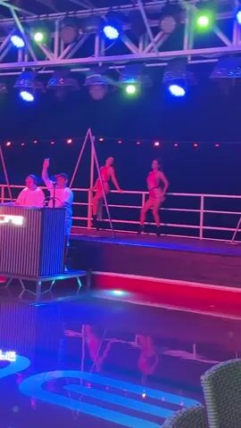 The video shows a stage with two dancers performing. The dancers are wearing what appears to be revealing outfits. A DJ is visible on the left side of the stage, and there is a triangular structure on the stage behind the dancers. The stage is lit with changing colored lights, including blue and purple. There are lights overhead and some red decorative lights strung along a railing. The floor in front of the stage appears to be wet, possibly a pool or a reflective surface, and the lights are reflecting off of it. The video is shaky at times.