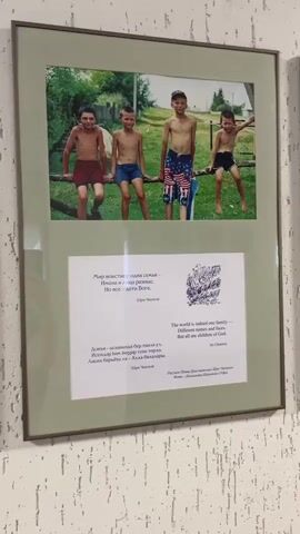 The video shows a series of framed photographs hanging on a wall with a light-colored patterned wallpaper. The first photograph shows four boys, shirtless and wearing shorts, sitting on a log or beam. Beneath the photo is a text in both Russian and English that reads "The world is indeed one family - different names and faces, But all are children of God." The second photograph shows two children, a boy and a girl, embracing and smiling. The text beneath this photo reads "The children of happiness will create a new world." The following two photographs and accompanying texts are not fully visible in the video. The overall impression is that the photographs are part of a display or exhibit related to children and global unity.