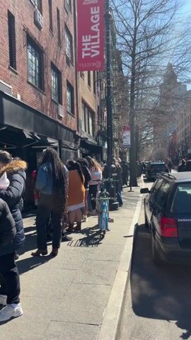 Here is a description of the video:

The video shows a long line of people waiting outside of a store on a sunny day. The line stretches down the sidewalk, with a mix of individuals of varying ages and clothing styles. A blue bicycle is parked near the front of the line. A black car is parked along the curb on the right side of the frame. There are buildings on both sides of the street, and a banner reading "THE VILLAGE CULTURE" is visible on a lamppost.
