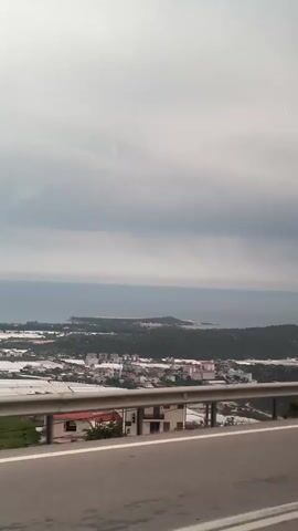 The video shows a view from a moving vehicle, likely a car, traveling on a road with a guardrail. In the background, there is a coastal landscape with buildings, greenery, and the ocean under a cloudy sky. The scene appears overcast, and the focus is on the vista beyond the road.