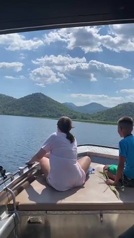 This video shows two children sitting on the back of a boat, looking out at a picturesque lake surrounded by lush green mountains. The sun is shining brightly, and the sky is dotted with fluffy white clouds, indicating it's a beautiful, clear day. The water is calm and blue, reflecting the sky and mountains.

The older child, a girl, is sitting cross-legged with her back to the camera, wearing a light pink t-shirt and pink shorts. She is holding onto the edge of the boat. A young boy in a blue t-shirt and green shorts is sitting beside her, facing slightly towards the camera. He is holding some green leaves in his hand. They appear to be enjoying a peaceful boat ride and taking in the scenic views.

The setting is a lake in Rogani, Montenegro, characterized by its serene natural beauty with rolling hills covered in vegetation. The time of day is clearly daytime, likely late morning or early afternoon, given the strong sunlight and clear sky. The overall mood conveyed is one of tranquility and enjoyment of nature. There are no visible text elements in the video.