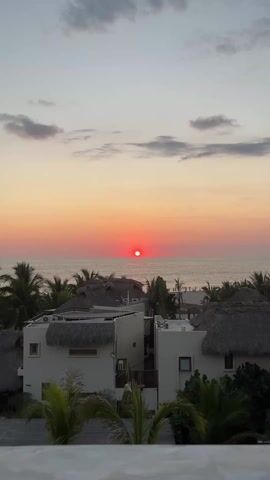 The video captures a sunset over the ocean, framed by beachside buildings with thatched roofs and palm trees. The sun is a bright, almost fiery red, positioned just above the horizon, casting a warm glow on the water and sky. A few people can be seen on the beach in the distance. The buildings in the foreground are white, and the scene gives a sense of tranquility and relaxation.