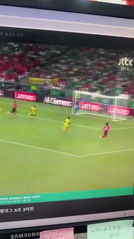 The video shows a computer screen displaying a soccer match. The screen is angled and reflects the surrounding environment. On the screen, there are players in yellow and red jerseys on a green soccer field with a goal in the background. A crowd is visible in the stadium seats, mostly in red. The JTBC LIVE logo is visible in the top right corner of the screen. Below the video, text is displayed, indicating it's a live broadcast of a soccer game. The lower part of the screen shows the bezel of a computer monitor, and some sticky notes are attached to it. One note has handwritten Korean text and a phone number.