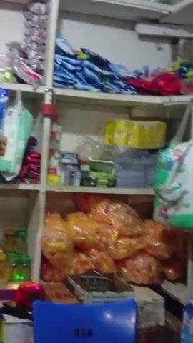 The camera pans over shelves in a small store, stocked with a variety of items. We see toiletries, food items, and other household goods. Some items are packaged, while others are loose in jars or containers. The shelves are white, and the lighting is dim.