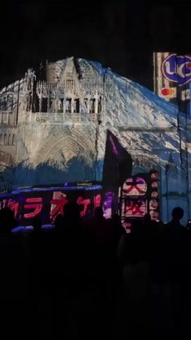 A video shows a nighttime scene with a large crowd of people silhouetted in the foreground, facing a towering building that is illuminated with colorful projections. The projections depict what appears to be a vibrant, neon-lit Japanese street scene with various signs and text, including "カラオケ" (Karaoke) prominently displayed. The building itself has intricate architectural details, suggesting it might be a cathedral or a similar historic structure, which is being transformed by the light show. The overall atmosphere is dark, with the focus on the brightly lit projections on the building. The camera remains static throughout the short clip, capturing the dynamic light display.