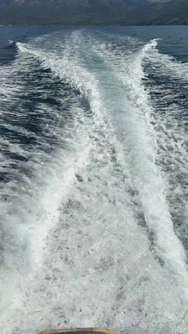 The video shows a view from the back of a boat traveling on the water. A prominent white wake trails behind the boat, expanding into a foamy churn. The water is dark blue on either side of the wake. In the distance, a coastline with green trees and a large mountain range under a clear sky is visible. The camera angle is looking directly behind the boat.