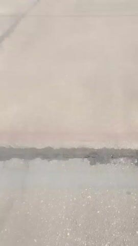 The video shows people walking on an airport tarmac. A white airplane is visible on the left. A man in a safety vest stands to the right. Others are walking toward the plane. The weather appears hazy or dusty. The ground is concrete with visible lines.