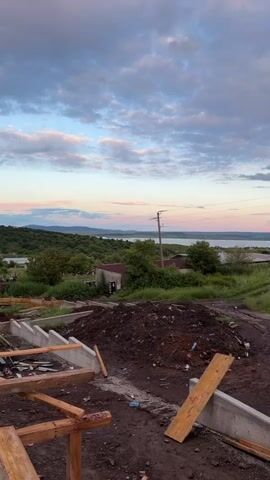 The video pans from left to right, showing a construction site with unfinished wooden frames and concrete foundations in the foreground. Behind the construction site, there is a dirt pile. The land slopes down into a grassy field, with a few buildings scattered throughout. In the background, there is a body of water stretching to the horizon, with a light pink and blue sunset visible above. Power lines run across the middle ground of the scene. The sky is mostly covered in clouds.