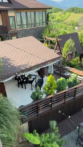 The video pans over a resort area nestled in a mountainous landscape. In the foreground, a wooden deck and patio area with gravel and decorative stones features outdoor seating, including a dining set with black chairs and a table, a barbecue grill, and a circular arrangement of lounge chairs around a fire pit. Several small evergreen trees and bushes are planted around the patio. Beyond the patio, several triangular-shaped houses with brown roofs are visible, surrounded by lush greenery. In the background, rolling hills covered in trees and terraced fields stretch towards the horizon, with layers of mountains visible in the distance under a cloudy sky. A larger building with large windows is also visible on an upper level. The overall impression is a scenic and tranquil resort with ample outdoor amenities. No people are visible in the video.