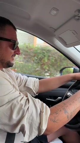 The video shows the interior of a car from two perspectives. The first perspective, from 00:00 to 00:01, shows a man with sunglasses and tattoos on his arm driving the car. The second perspective, from 00:01 to 00:04, shows the car's dashboard with a navigation screen displaying a map, and the road ahead. The car is driving down a street lined with buildings, parked cars, and trees. The sky is overcast.