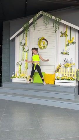 A young girl with her hair in pigtails is performing a split on a step in front of a decorative white structure resembling a house facade. The structure is adorned with spring-themed decorations, including wreaths, ribbons, and yellow flowers. The girl is wearing a neon green crop top and black leggings. She is in the middle of a split, with her legs extended to the sides. She waves to the camera with her right hand. The setting appears to be outdoors, possibly on a stage or elevated platform, with a tiled floor in the foreground. The overall mood is cheerful and festive, likely related to a spring or Easter celebration.