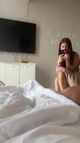 A woman is sitting on a bed looking at her phone. She is wearing a green tank top and has long brown hair. She is holding a pillow in her lap. Behind her is a wall with the words "С ДНЕМ РОЖДЕНИЯ" written in large white letters. To the left of the woman is a white dresser with a TV on top of it. In front of her is a bed with white sheets. The lighting in the room is bright.