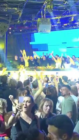 The video shows a crowded nightclub with a DJ performing on stage. The stage has a large blue screen displaying "TERMINAL". The crowd is enthusiastically dancing with their arms raised, some holding phones to record. The lighting is bright, with spotlights shining onto the stage and the crowd. The atmosphere is energetic and festive.