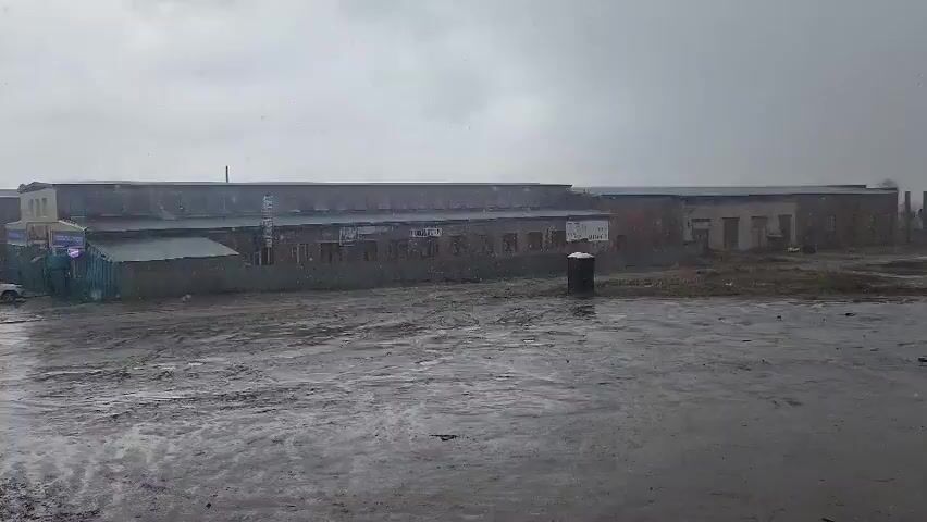 The video shows an outdoor scene in an industrial or semi-industrial area under a heavily overcast, gloomy sky. Snow or sleet is falling, visible as white specks against the dark sky and buildings. In the background, there are several long, low-rise brick and concrete buildings that appear to be warehouses or factory structures. One building on the left has "VINTAZH" (VINTAGE) written on its side. The foreground consists of a large, muddy, and wet expanse of ground, with puddles reflecting the dull light. A black bin or barrel stands in the middle of this muddy area. Briefly, a white car drives from the left side of the frame, partially obscured by other structures, before disappearing to the right. The overall impression is one of a cold, wet, and somewhat desolate industrial landscape during daytime.