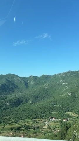 The video shows a roadside view of rolling green mountains under a clear blue sky. In the foreground, a metal railing is visible. To the left, a large billboard displaying an image of a building and text is partially visible. The overall scene is one of a scenic natural landscape.