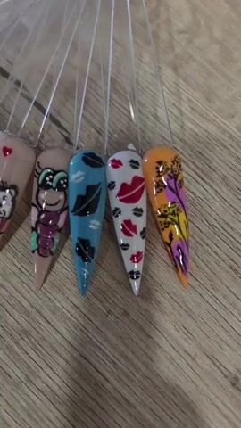 A fan of artificial fingernails is displayed on a wooden surface. The fingernails feature various designs, including cartoon characters, lips, floral patterns, and animal prints. The lighting is bright, illuminating the detailed artwork on each nail. The video pans across the collection, offering a clear view of the different styles and colors.