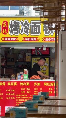 The video shows a food stall in China. A man in a black t-shirt is working inside the stall, presumably preparing food. The stall has a bright yellow sign with Chinese characters. The sign also mentions "fresh ingredients" and "rich sauce." The menu, displayed in red, lists various items and prices, including "grilled cold noodles." There are several small boxes of tissues on a wooden table in front of the stall. The environment appears to be a busy street or market.