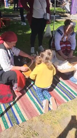 Here is a concise description of the video:

The video shows two women in traditional Romanian clothing sitting on a colorful striped rug. They are teaching two young children to work with wood, possibly carving or shaping it with tools. They sit in a grassy area with other people standing around. The area is possibly an outdoor market or cultural event. One of the women is wearing a red headscarf and the other is wearing a white blouse with embroidery. The children appear to be enjoying the activity.