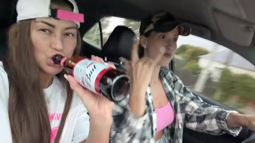 Two women are in a car. The woman in the foreground is wearing a white t-shirt and a white baseball cap with a pink strap. She is holding a bottle of Budweiser beer and drinking from it. Her hair is long and brown. The woman in the background is wearing a plaid shirt over a pink top and a camouflage baseball cap. She appears to be driving. The scene is blurred, suggesting the car is in motion, likely during the daytime, given the natural light. The background shows a residential area with houses and trees, suggesting a suburban or rural setting. The mood appears casual and lighthearted, with the women seemingly enjoying themselves while driving. There is no visible text in the frame, other than the branding on the beer bottle.