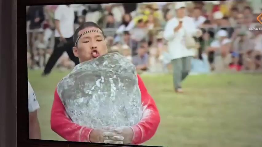 A man in a red shirt and a headpiece struggles to carry a large, rough gray rock. His hands are clasped around it, and his face is strained with effort. He appears to be in an outdoor arena with a crowd of spectators in the background. Some spectators are seated, while others stand. The ground is covered in grass. The scene is filmed from a medium distance, capturing the man and his immediate surroundings. There's a blurred logo in the upper right corner of the screen.
