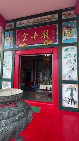 The video shows the exterior and interior of a small Chinese temple. The temple's entrance is painted red and framed with dark green panels featuring images of cranes, people, and decorative motifs. Above the entrance, a red sign with golden Chinese characters is visible. Inside, there are statues on a decorated altar, flanked by pillars adorned with dragons.  Cushions are laid on the floor for worshippers. The temple's interior is lit by a chandelier.