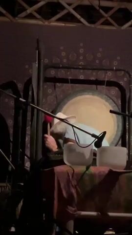 A person wearing a light-colored hat and blue shirt is playing a large gong with a red mallet. The gong is suspended in a dark frame, and microphones are positioned in front of it. In the foreground, two crystal bowls are visible, resting on a colorful cloth. The background features a patterned tapestry or curtain. The scene appears to be indoors, possibly a performance space or meditation room, with stage lighting visible at the top of the frame. The overall atmosphere is serene and focused on the musical performance. The timestamp "00:00" and "00:05" are visible in the video. The location context is ตำบลเกาะพะงัน, Thailand.