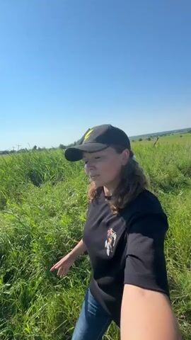 A person wearing a black t-shirt and a black baseball cap with a yellow logo is standing in a grassy field under a clear blue sky. The person is looking off to the side and appears to be holding a camera or phone to record a video. The field is lush with tall green grass.