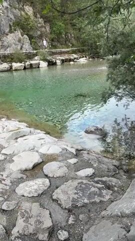 The video opens with a wide shot of a clear, turquoise river flowing over a rocky bed. Large, gray stones line the foreground, some submerged in the water. In the middle ground, a low, man-made weir causes a section of the river to cascade over, creating white froth. The water is exceptionally clear, allowing the rocky bottom to be seen.

As the video progresses, the camera pans to the right, following the curve of the river. The left bank is steep and rocky, covered in sparse vegetation. In the distance, a person in white clothing stands on a paved path that runs along the riverbank. Trees and dense green foliage line the top of the rocky bank.

The camera then moves closer to the weir, showing the water spilling over its concrete structure. The green hue of the water intensifies in this area, possibly due to algae or the depth of the pool. The sunlight reflects off the water's surface, creating shimmering patterns.

Further to the right, a concrete structure, possibly part of a small building or pumping station, is visible at the edge of the water, near the weir. The overall environment appears to be a natural, mountainous landscape with a significant watercourse. The video concludes with a closer view of the weir and the vibrant green pool below.