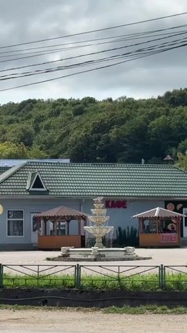 A multi-tiered water fountain is in the foreground of a single-story building with a green tiled roof. The building has a sign that reads "КАФЕ" in red letters. To the left and right of the fountain are two open-air gazebos with brown roofs. In front of the building, there is a paved area and a low fence. Behind the building, a forested hill rises. Overhead, power lines are visible against a cloudy sky. A dark-colored car drives past in the bottom left corner of the frame.