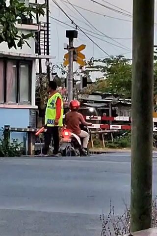 The video shows a man in a bright yellow vest and a red shirt standing next to a motorcycle with a rider wearing a red helmet. The man in the vest is holding a bright orange object, possibly a signal bat. They are positioned at a railroad crossing, with the crossing gate down, indicating that a train is approaching or passing. The train is visible in motion, a long gray and orange vehicle. The environment appears to be outdoors with trees and power lines visible. The overall scene suggests a traffic controller or railway worker managing the crossing during a train passage.
