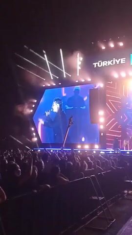 The video captures a live concert performance in an outdoor setting. A large screen displays a singer in a black suit, holding a microphone and standing next to a string instrument on stage. The stage is illuminated with bright white spotlights and colorful LED lights, and there is smoke or haze in the air. In front of the stage, a large crowd is visible, with many people raising their hands and cheering. A metal barricade separates the crowd from the stage area. The words "TÜRKİYE KÜLTÜR YOLU FESTİVALİ" are displayed on the screen above the stage. The overall atmosphere suggests an energetic and well-attended event.