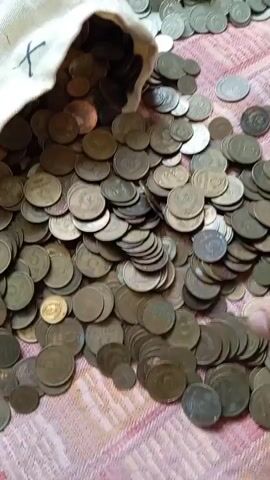 A hand is pouring a pile of coins from a beige fabric bag onto a patterned surface. The coins are a mix of bronze and silver-colored metal, with some appearing to be older and worn. The surface is a pink and white textured material. The hand is large and appears to be male. The coins are falling in a cascade, creating a dense layer on the surface. The video is shot from a slightly elevated angle, focusing on the coins and the hand.