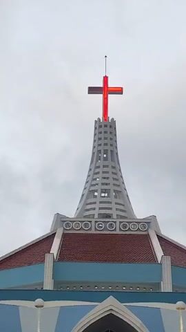 A vertical video shows the top of a church building with a red cross illuminated at the apex of a tall, white, tower-like structure. The tower has a lattice-like design with horizontal bands of open spaces. Below the tower is the domed roof of the church, which has red shingles and a light blue band encircling it. Around the base of the tower, on the roof, are circular decorative elements with what appear to be letters within them, forming the word VERITA. The sky is overcast and gray. The video is 4 seconds long and has no discernible audio.