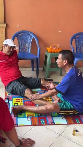 Two men are sitting on the floor. One man, wearing a red shirt and a white cap, is seated with his legs extended. The other man, wearing a blue shirt and shorts, is kneeling in front of him and massaging his leg. They are on a colorful mat in front of an orange wall, with blue chairs and a green shelf in the background. There is also a small bottle of oil on the floor near the mat.
