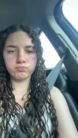 A young woman with curly brown hair and a white tank top is seated in a car, wearing a seatbelt. She makes a kissing face at the camera. The interior of the car is visible, including the door frame and black leather seats. The window shows some trees passing by. The woman then shows her left hand displaying a peace sign. Her nails are long and painted with a white base and blue tips.