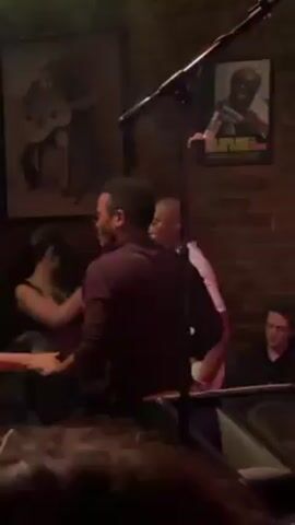 A group of people are dancing in what appears to be a small, dimly lit bar or club.  Several people are moving to music. Some have their hands outstretched as if partnered in a dance. The setting includes exposed brick walls decorated with framed pictures, and string lights across the ceiling. Several audience members are visible, watching the dancers.
