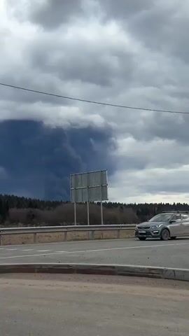 A video taken from the side of a road shows a massive, dark column of smoke rising into an overcast sky. The smoke plume is very dense and appears almost black against the cloudy gray sky, towering over a line of trees in the distance. The scene suggests a large fire or industrial incident beyond the visible forest.

In the foreground, a gray Kia Rio car with roof rails drives past the camera from right to left. A person is visible in the driver's seat. The car has a Russian license plate, "M305ET 159". A metal guardrail runs alongside the road. The road surface is asphalt, and there's a dirt shoulder next to the guardrail. The trees forming the horizon appear bare or sparsely leaved, suggesting late winter or early spring. The sky is heavily clouded, contributing to the somber mood of the scene.