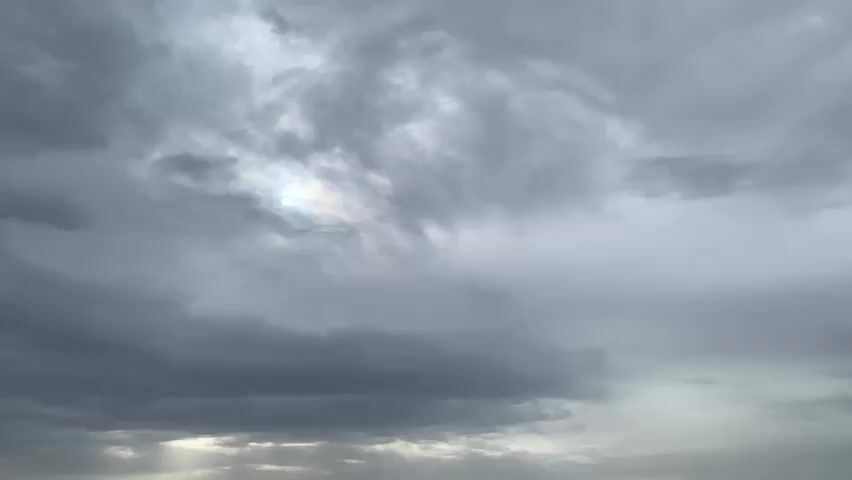 This media file shows a time-lapse of a cloudy sky over Kuru Karama, Nigeria. The clouds are predominantly gray and appear dense, suggesting overcast or possibly stormy weather. Sunlight occasionally peeks through breaks in the clouds, creating illuminated patches and shafts of light, particularly towards the bottom of the frame. The scene is natural and devoid of any specific man-made objects or human figures. The time of day is not explicitly indicated, but the lighting suggests it could be late afternoon or early morning, given the diffused sunlight. The overall mood conveyed is somber or dramatic due to the heavy cloud cover, yet the glimpses of light offer a subtle contrast. There is no visible text in the media file.