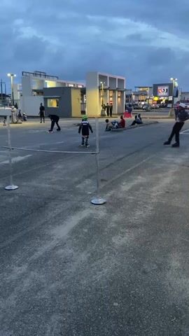 In a dusky outdoor setting, a group of people are roller skating, apparently engaging in a limbo contest. One individual in a white helmet, dark clothing, and roller skates successfully leans back and navigates under a horizontal pole supported by two vertical stands. There are other skaters and people scattered around the paved area. The backdrop features low-rise buildings, street lamps, and a sign that reads "House of Lekki" in bright colors. The sky is overcast, contributing to the dim lighting.